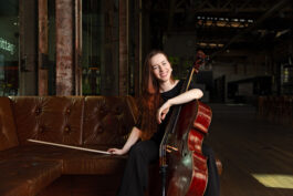  Anna Jeger sitzt auf dem braunen Sofa. linker Arm um das Cello gelegt, mit dem rechten Arm stützt sie sich auf dem Sofa ab. Der Cellobogen liegt da. Sie lacht in die Kamera.