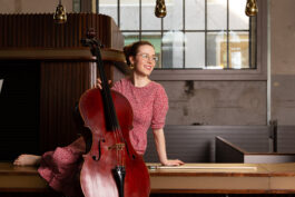 Anna Jeger sitzt seitwärts, im roten Kleid auf einem Thresen, hält ihr Cello und schaut lächelnd links an der Kamera vorbei.