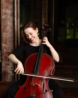  Anna Jeger spielt Cello und schaut lächelnd zu ihrer Bogenhand nach rechts unten. Der Hintergrund ist dunkel, die rote Farbe des Intruments leuchtet.
