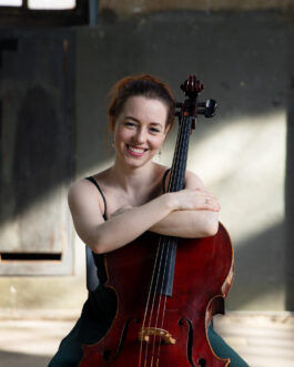 Portrait von Anna Jeger wie sie ihr Cello umarmt; sitzt auf Stuhl und schaut lachend in die Kamera.